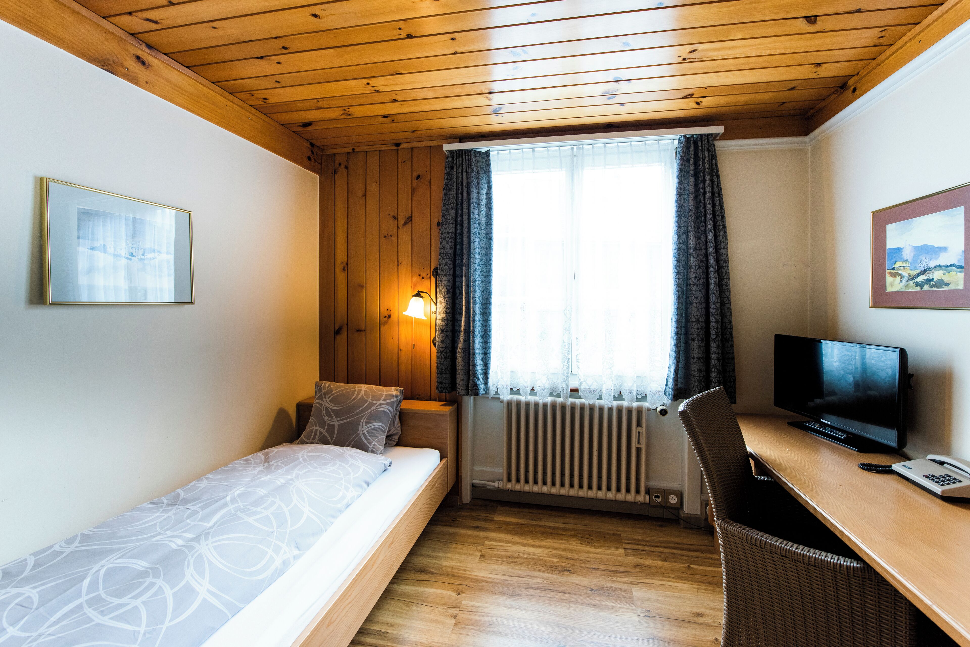 Una habitación pequeña y acogedora con techo de madera, cama con ropa de cama estampada, escritorio con computadora y ventana con cortinas.