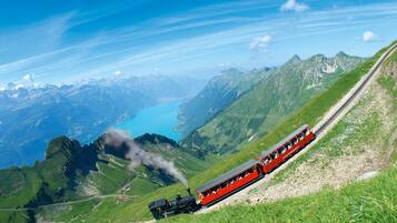 A red train on a mountainous track with a lake and mountains in the background.
