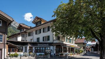Um hotel europeu tradicional com entrada central, área externa para sentar e montanhas ao fundo.