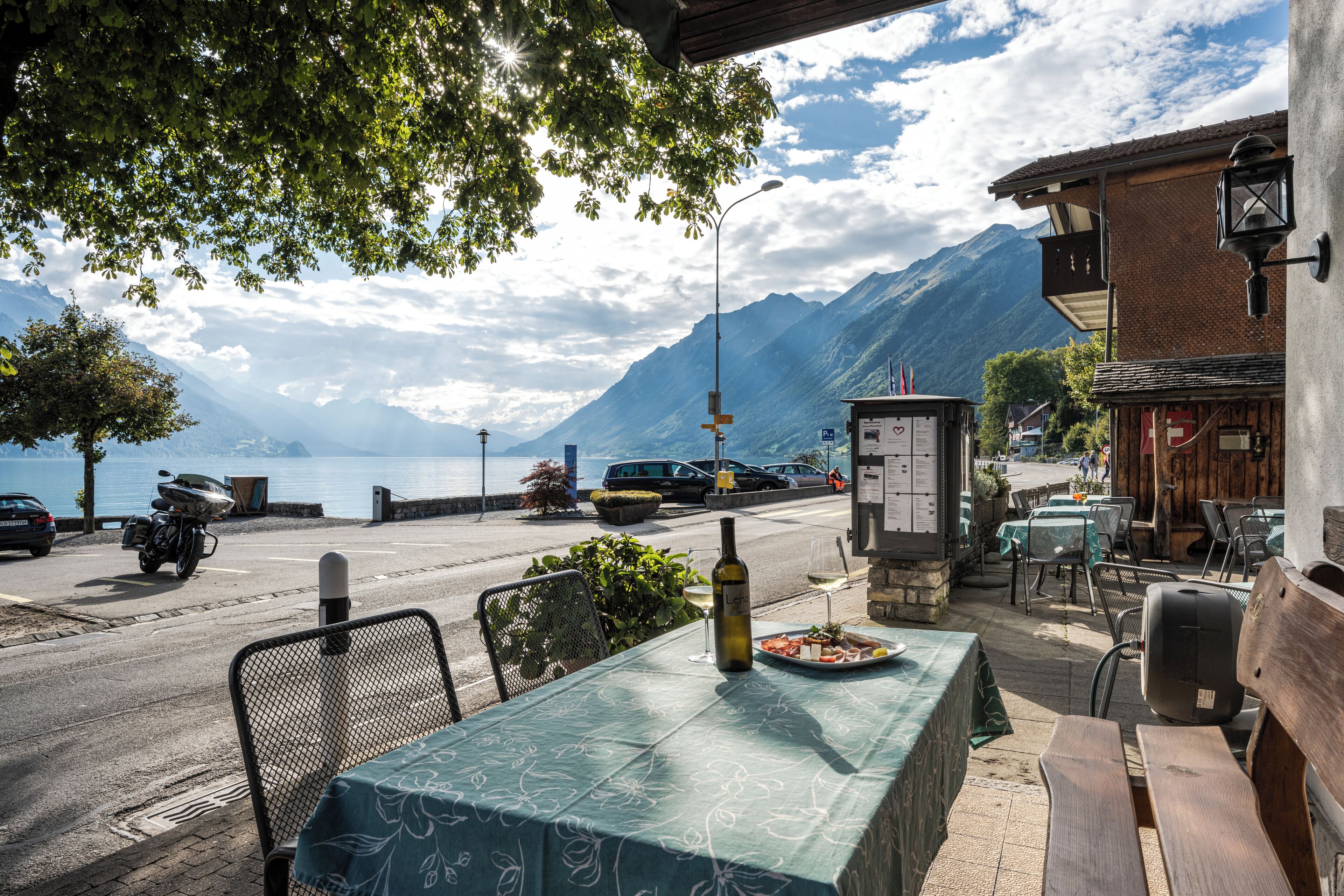 A table with a bottle and a plate of food, a chair, and a view of a mountain and lake.