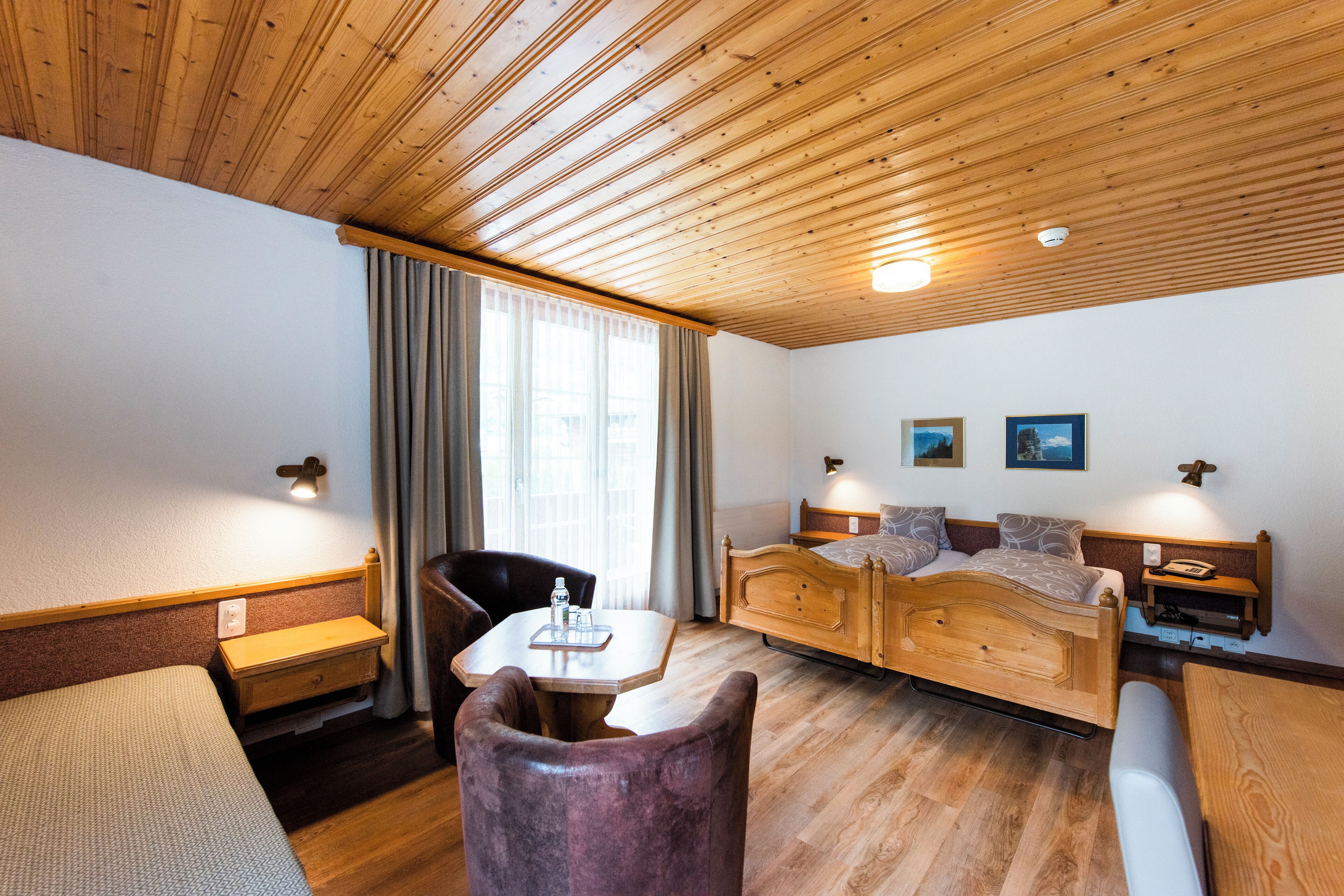 A room with a wooden ceiling, a bed, a desk, a chair, and a window with curtains.