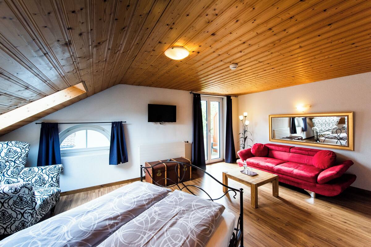 Una stanza accogliente con soffitto a falde, un divano rosso, un tavolo di legno, un letto con lenzuola decorate e una televisione, oltre a una finestra con tende.