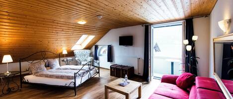 Un dormitorio con una cama, un sofá rojo, una mesa de centro de madera, una mesita de noche con una lámpara, una silla y un televisor en la pared.