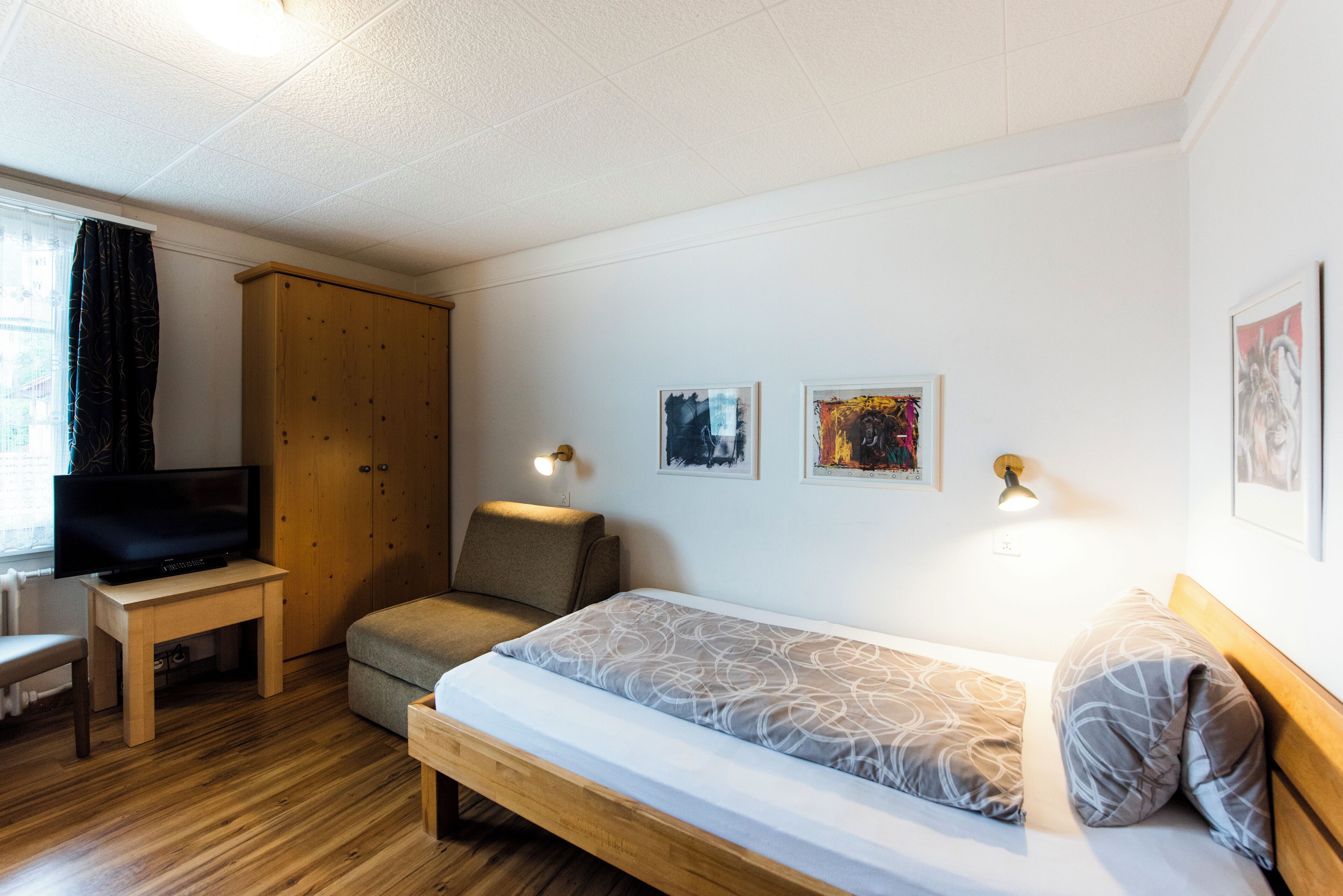 A bedroom with a bed, a TV, a chair, a wardrobe, and two wall-mounted lights.
