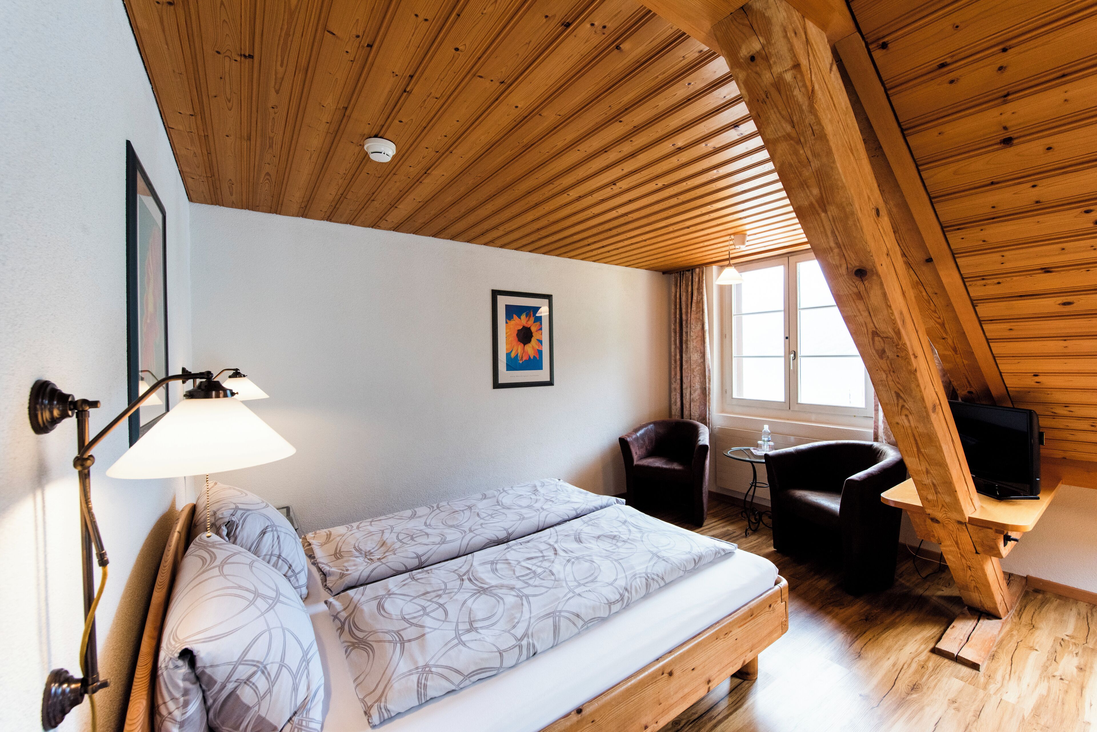 A bedroom with a wooden ceiling, a bed with patterned bedding, a desk with a television, and a chair.