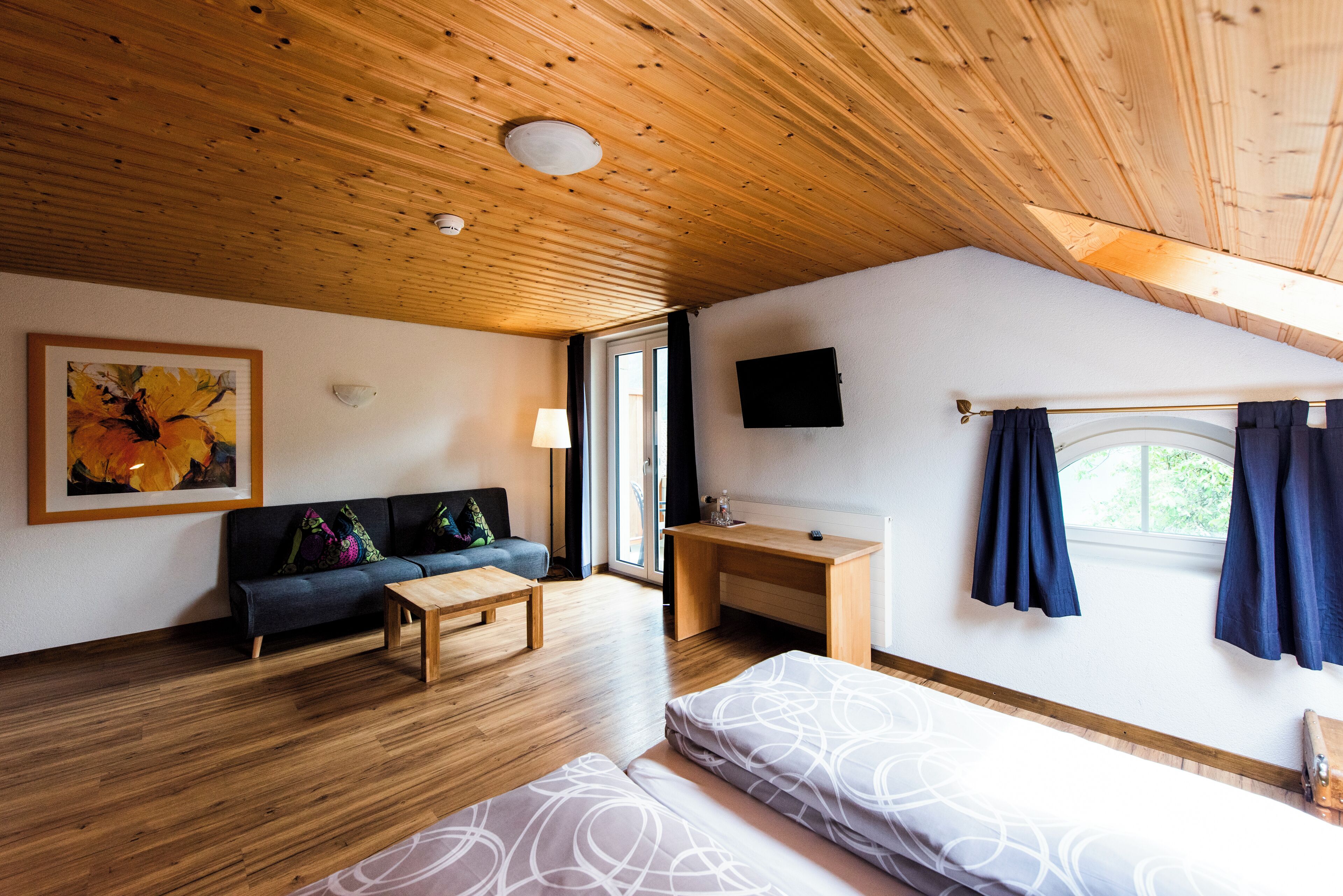 A room with a wooden ceiling, a sofa, a bed, a desk, a TV, and a window with curtains.