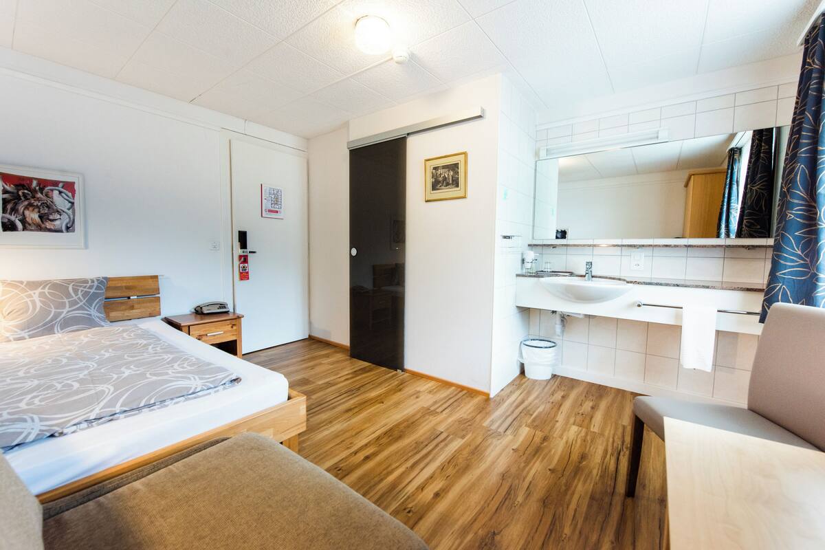 Una camera d'albergo moderna con pavimento in legno, un letto con copriletto decorato, un piccolo comodino, un tavolino notturno, un bagno con lavandino e specchio bianchi e una porta che dà su un'altra stanza.
