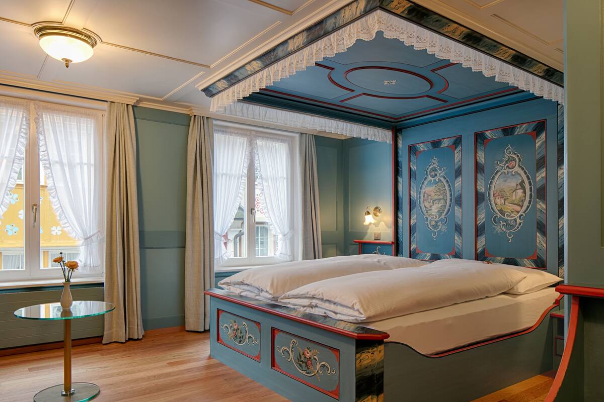Ein Schlafzimmer mit einem Bett, einem runden Glastisch mit einer Vase und einem Fenster mit Vorhängen.