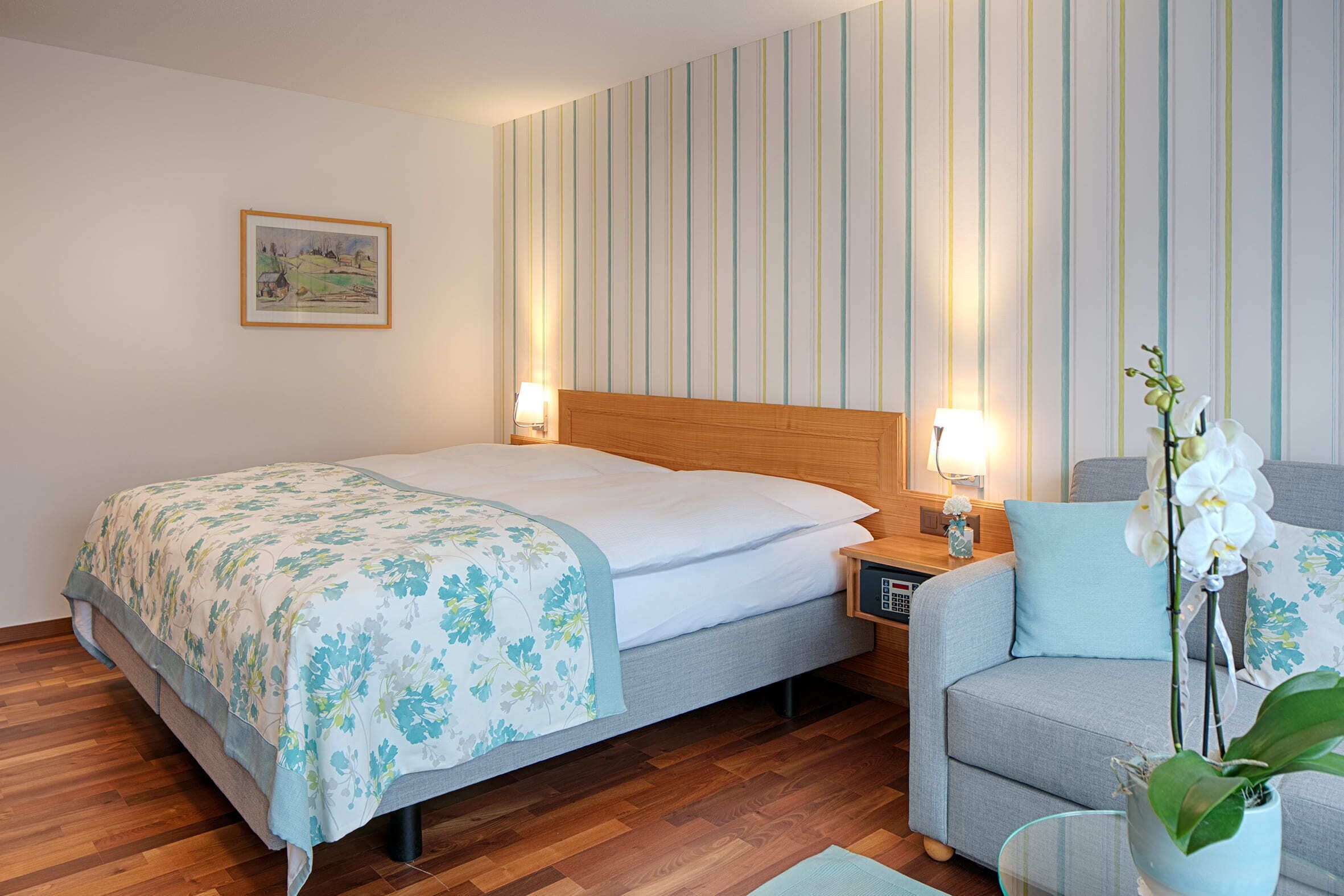 Una habitación de hotel con cama, mesita de noche, lámpara, silla y una orquídea en maceta.
