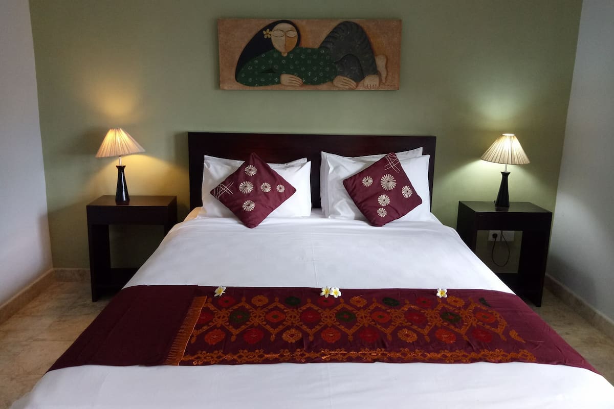 A neatly made bed with white linens, two decorative pillows, a patterned throw, and a painting above the headboard.