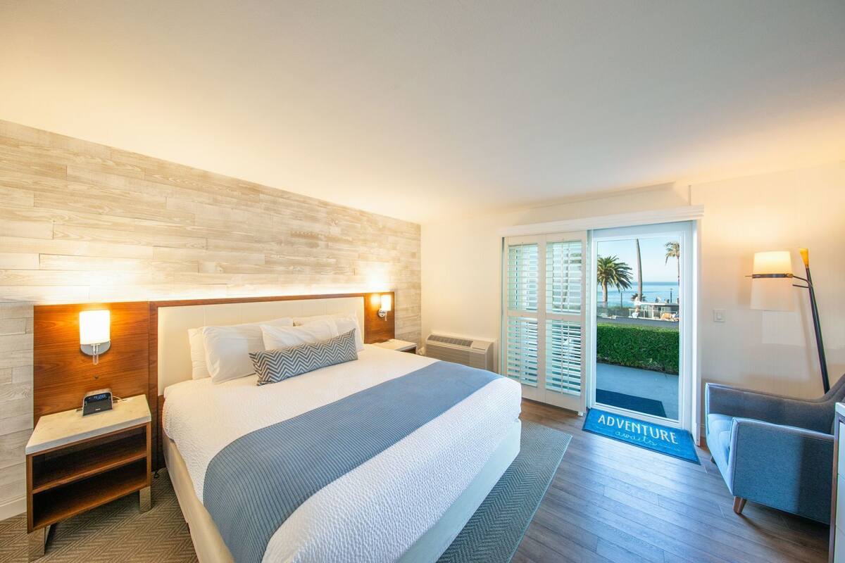 Habitación de hotel con cama, mesitas de noche, lámpara y vista a la playa por la puerta.