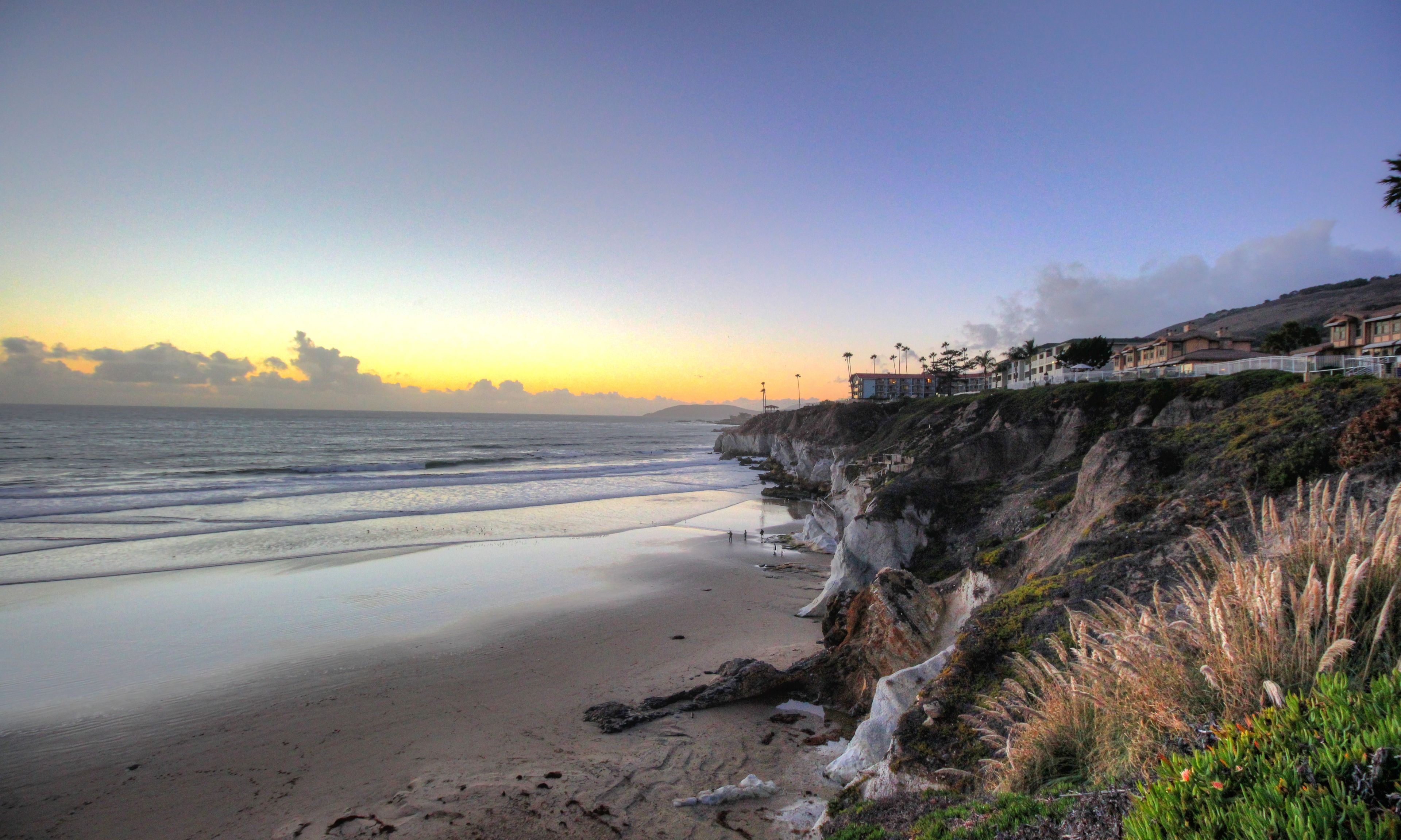 A coastal beach scene with a rocky cliff and a building with a balcony overlooking the ocean.