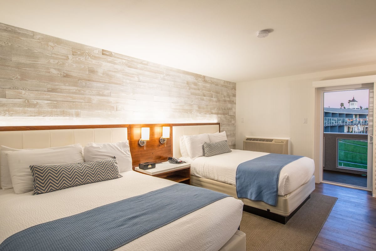 Habitación de hotel con dos camas, cabecero de madera, mesita de noche con teléfono y vista a un edificio a través de una puerta de vidrio.