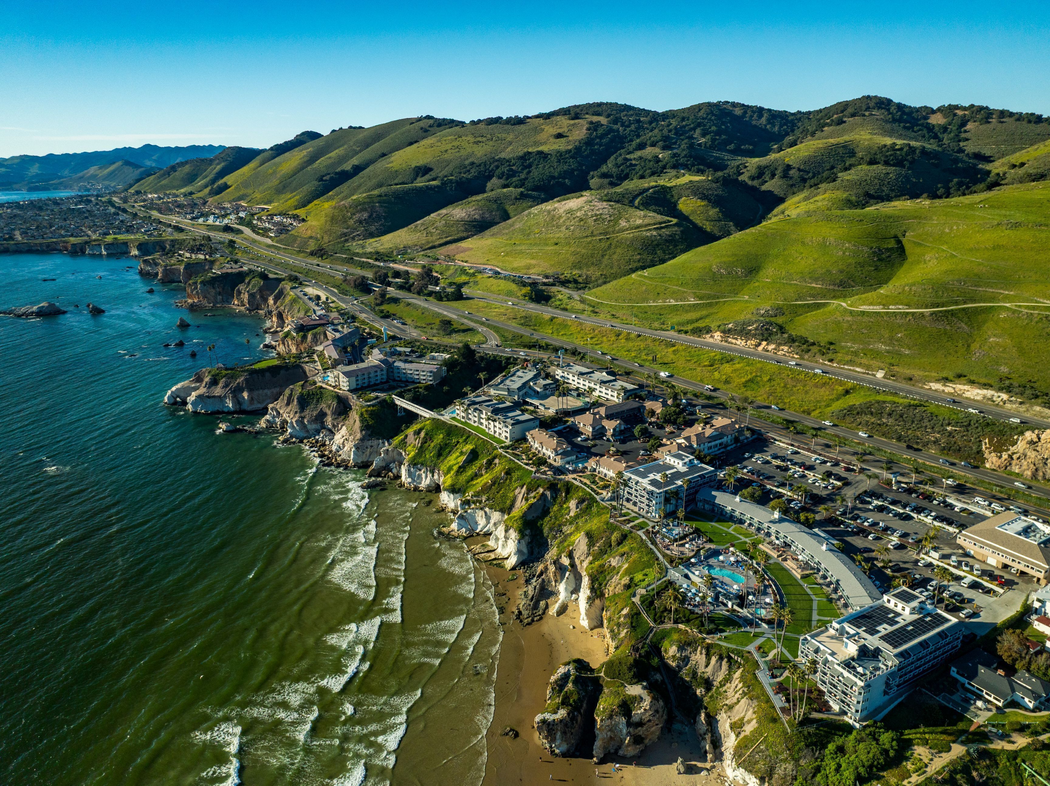 An aerial view of a coastal resort with a marina, beachfront, and hilly terrain in the background.