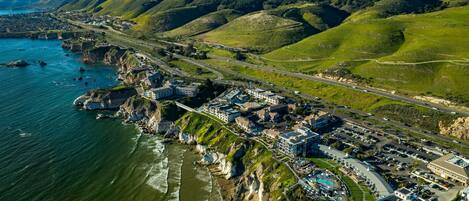 An aerial view of a coastal resort with a marina, beachfront, and hilly terrain in the background.