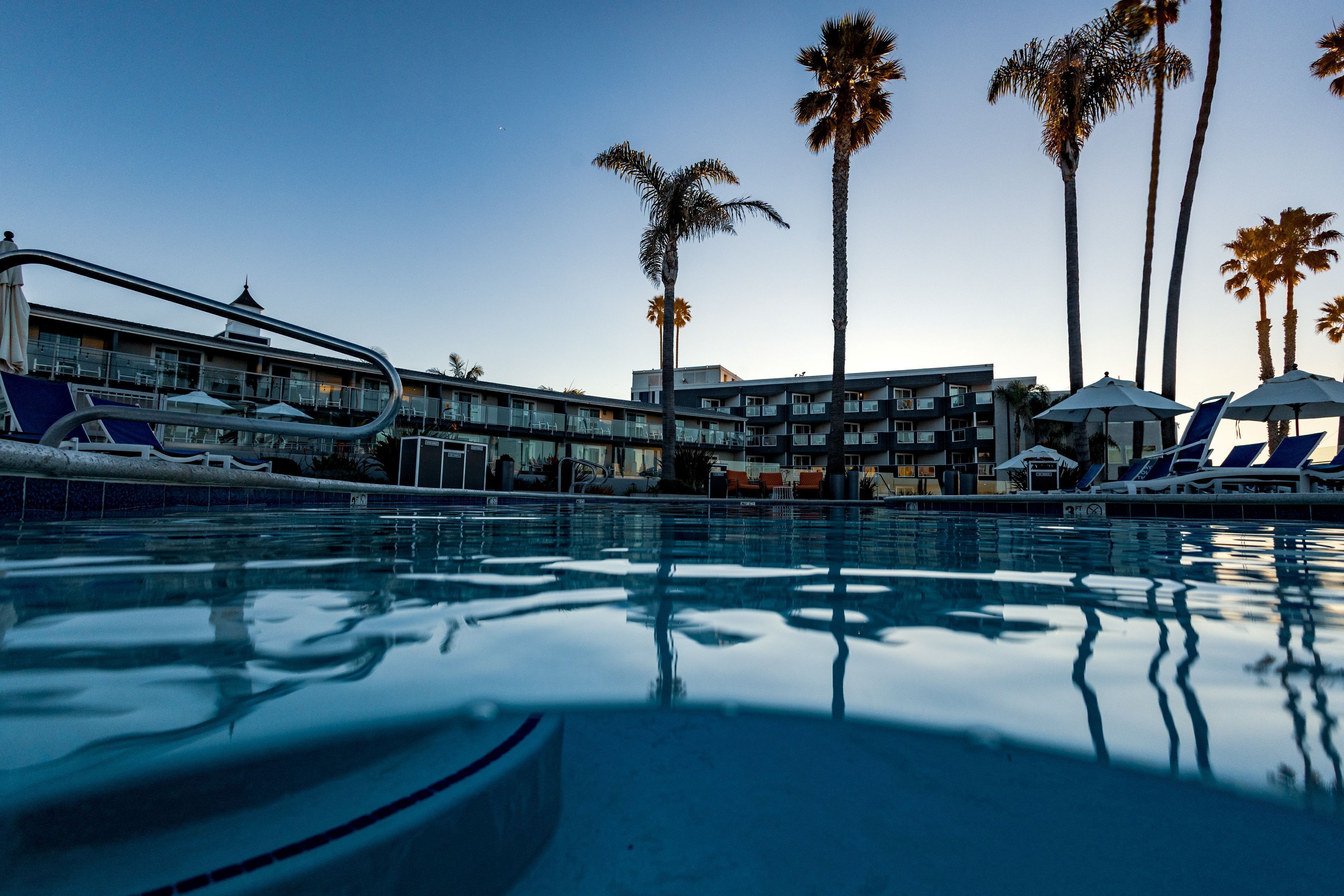A swimming pool with lounge chairs and umbrellas, surrounded by palm trees and a hotel building with balconies.