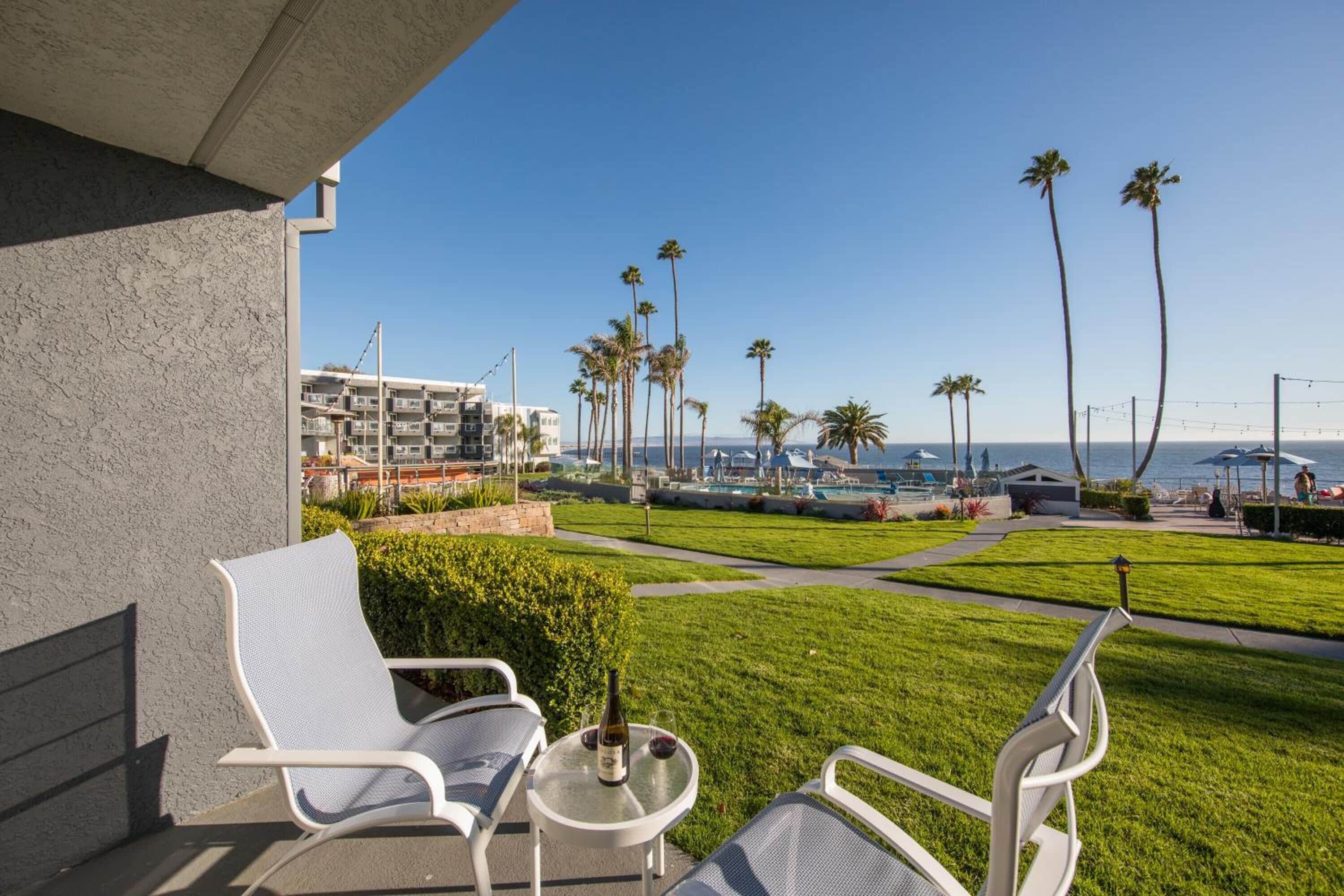 A balcony with two white chairs and a small table, overlooking a well-maintained lawn with palm trees and a view of the ocean.