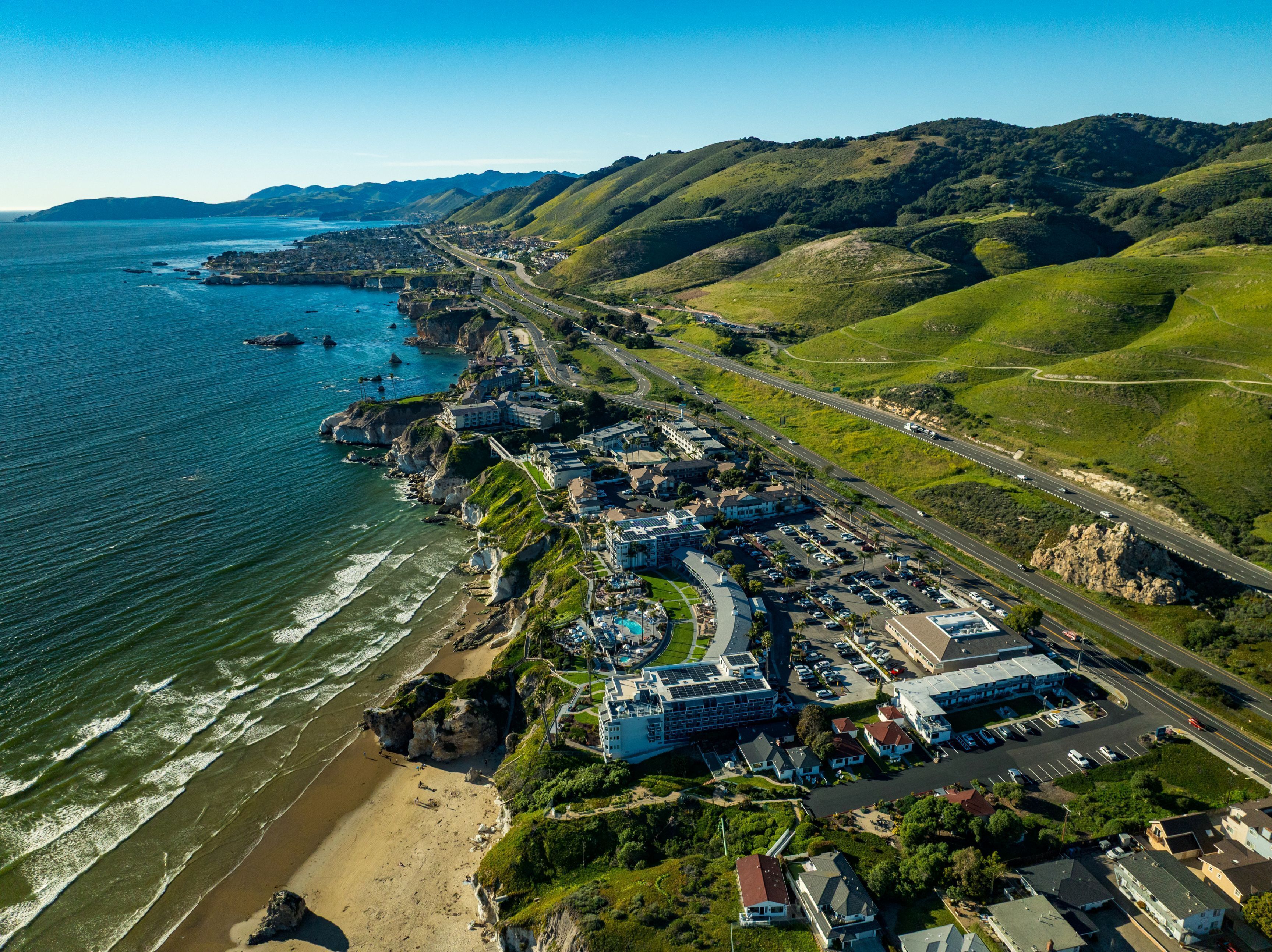 An aerial view of a coastal town with a beach, a marina, and a road leading to the town.