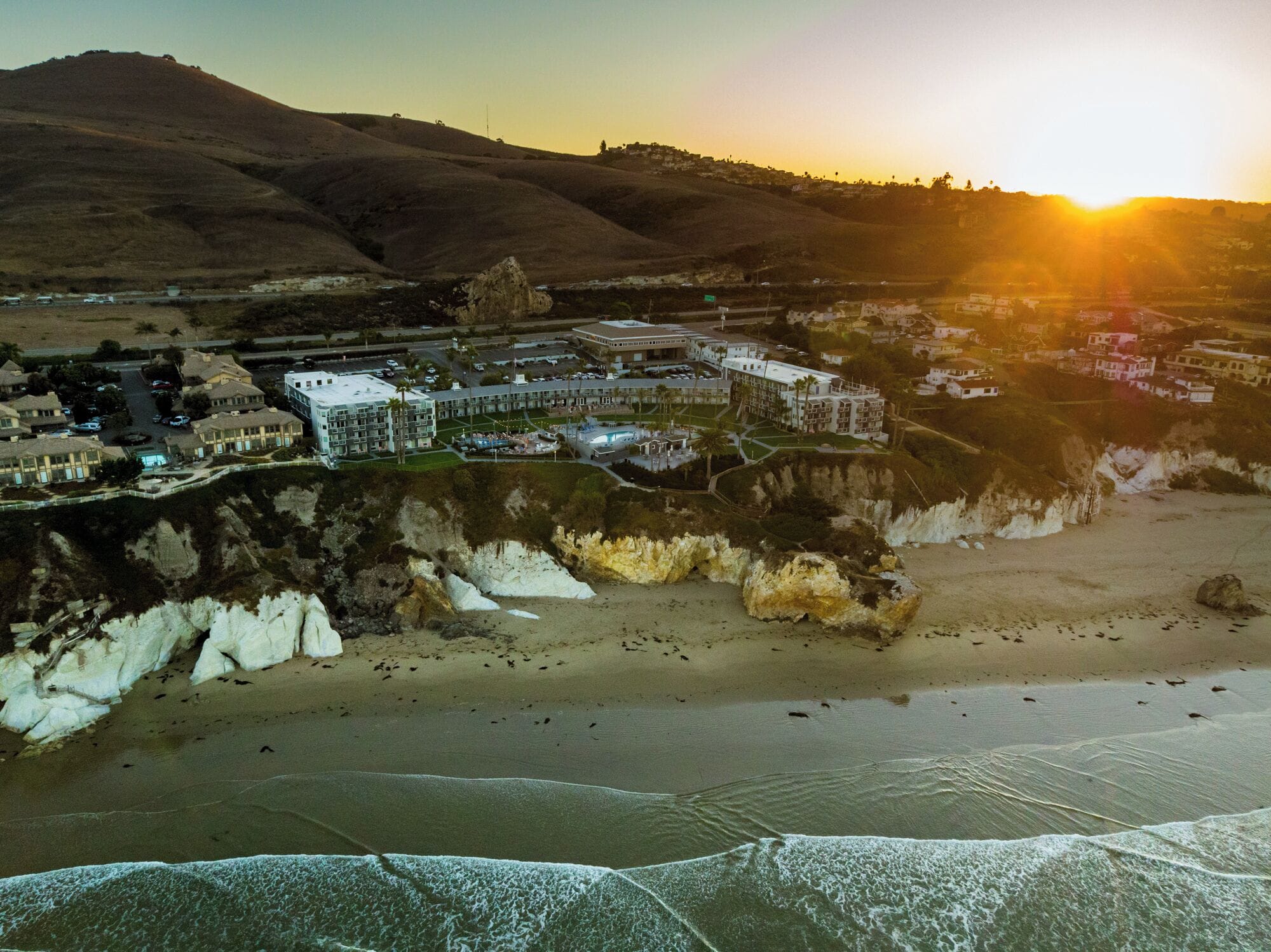 An aerial view of a coastal resort with a beachfront, a large hotel complex, and surrounding hills.
