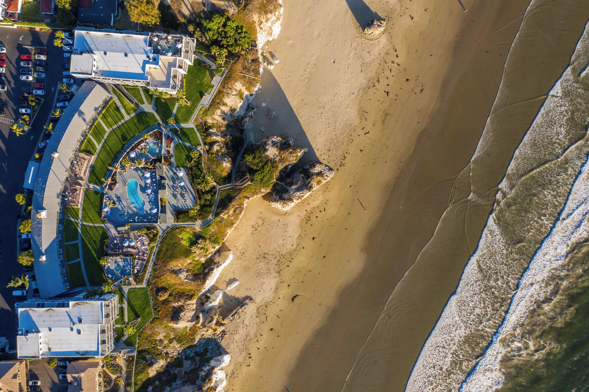 Vista aérea de un hotel costero con piscina, acceso a la playa y zona de estacionamiento.
