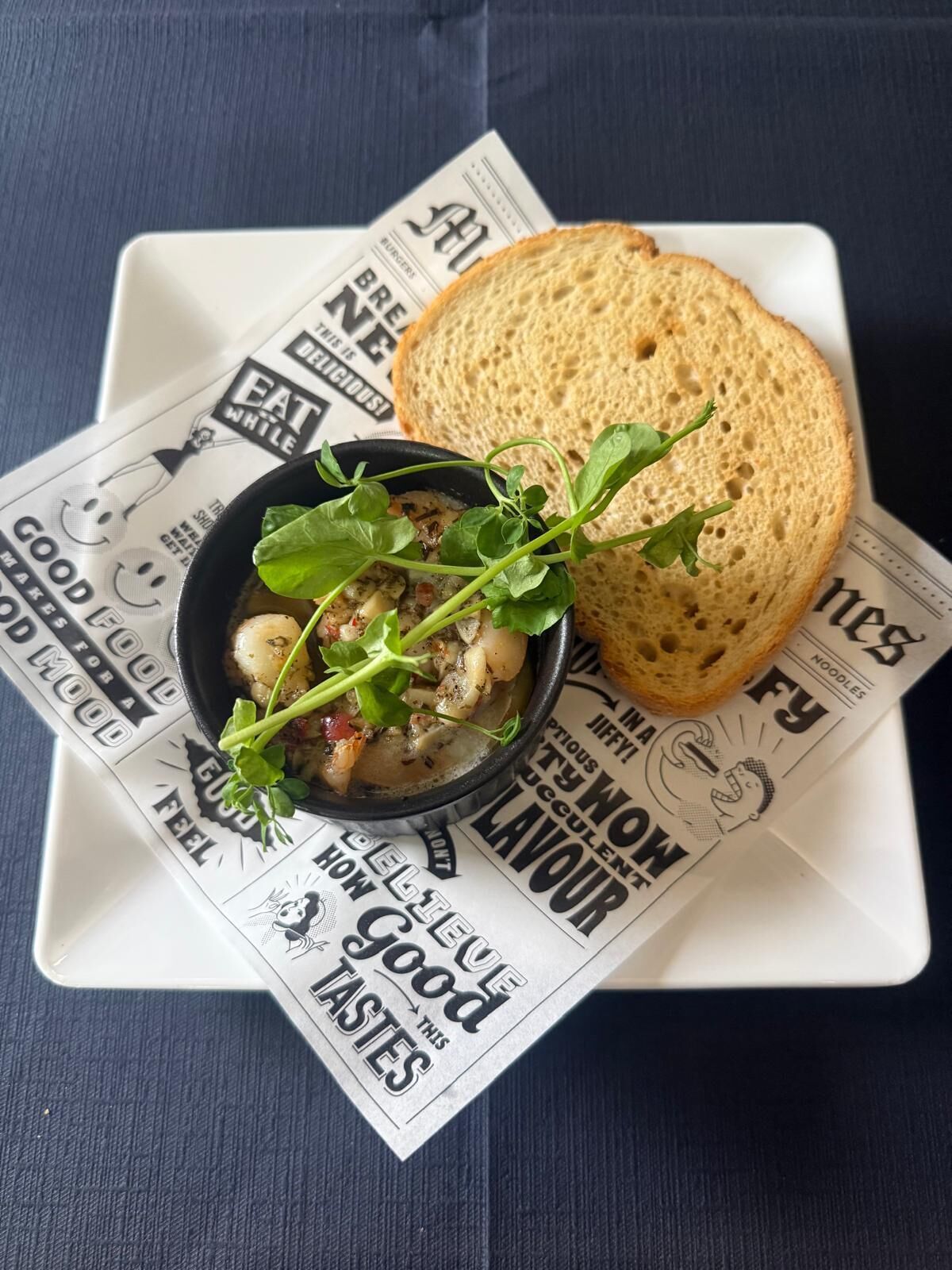 A plate with a bowl of food and a slice of bread on top, placed on a table with a newspaper underneath.