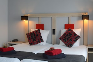Habitación de hotel con dos camas, cada una con almohada de diseño rojo y negro, ropa de cama blanca y cabecero de madera. Hay dos mesitas de noche, una con lámpara y teléfono.