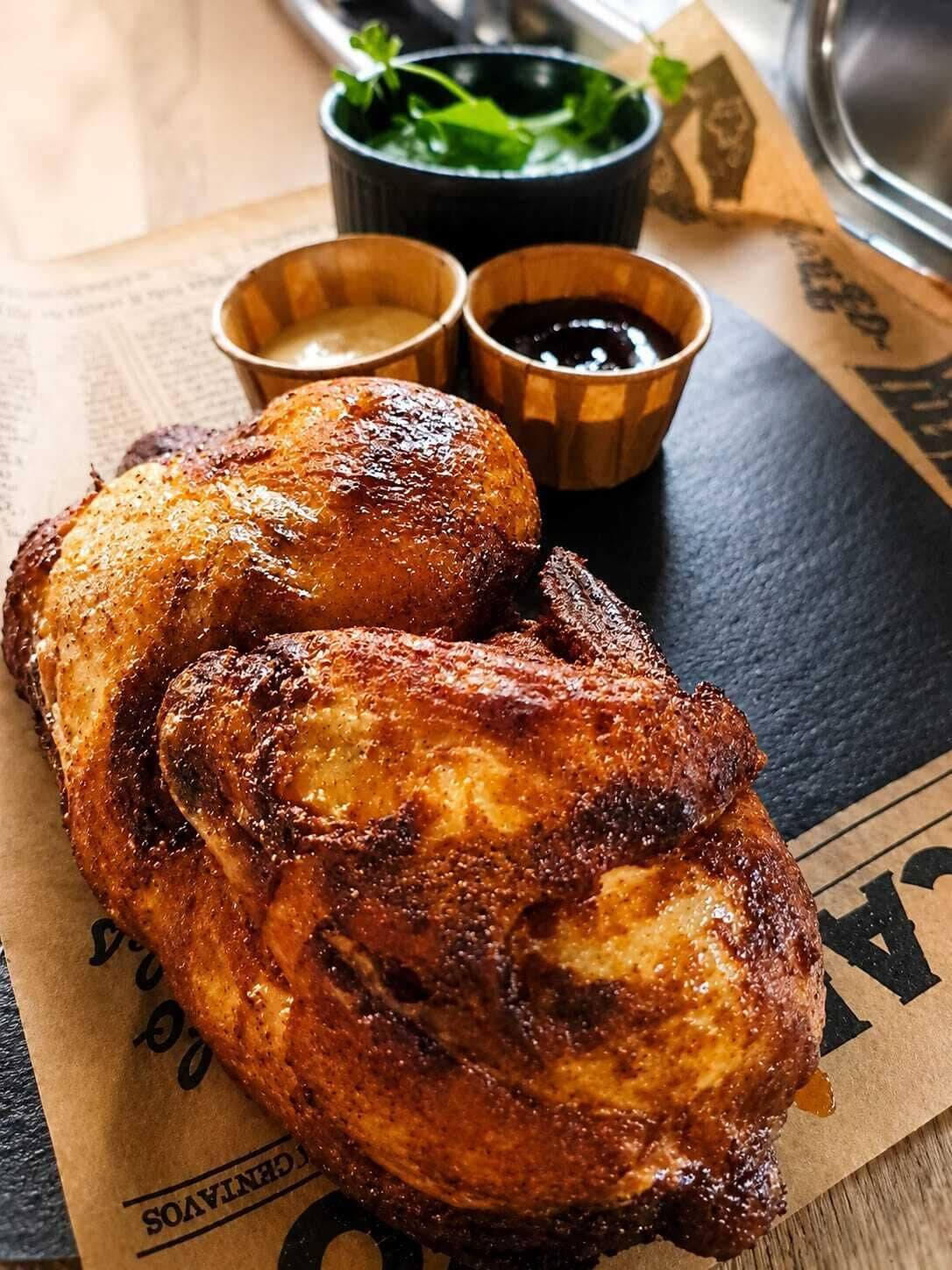 Un poulet rôti, accompagné de deux sauces pour tremper et d’une branche de persil, est posé sur une table.