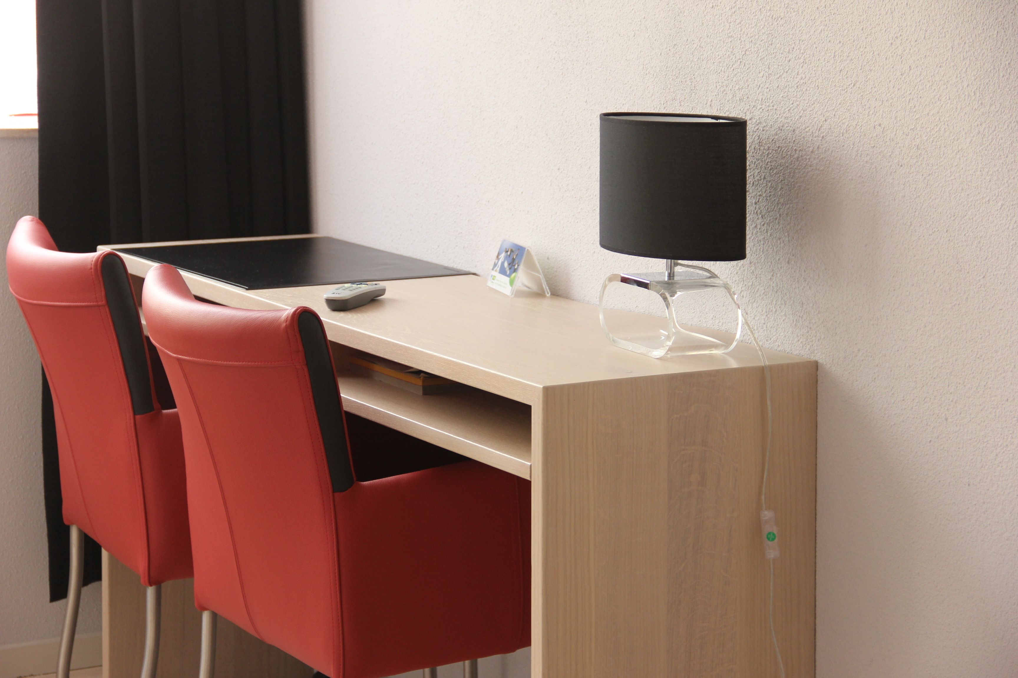 Habitación de hotel con escritorio, monitor de computadora, teléfono inteligente y sillas rojas.