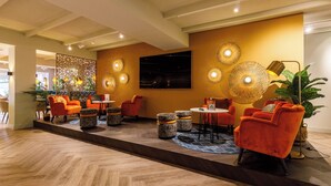 A modern lounge area with orange chairs, a large TV screen, decorative wall lights, and a patterned rug.