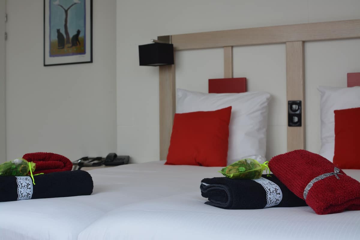 A hotel room with a neatly made bed, red and white pillows, and a painting on the wall.