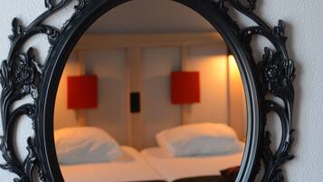 A mirror with an ornate black frame reflecting a hotel room with two beds and red wall lamps.