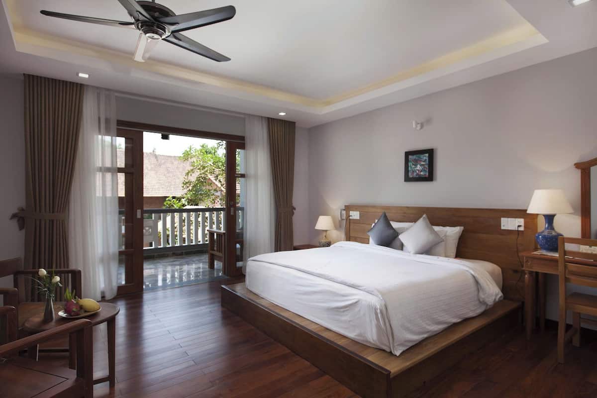 A hotel room with a large bed, wooden flooring, a ceiling fan, a balcony with a view, and a desk with a lamp.