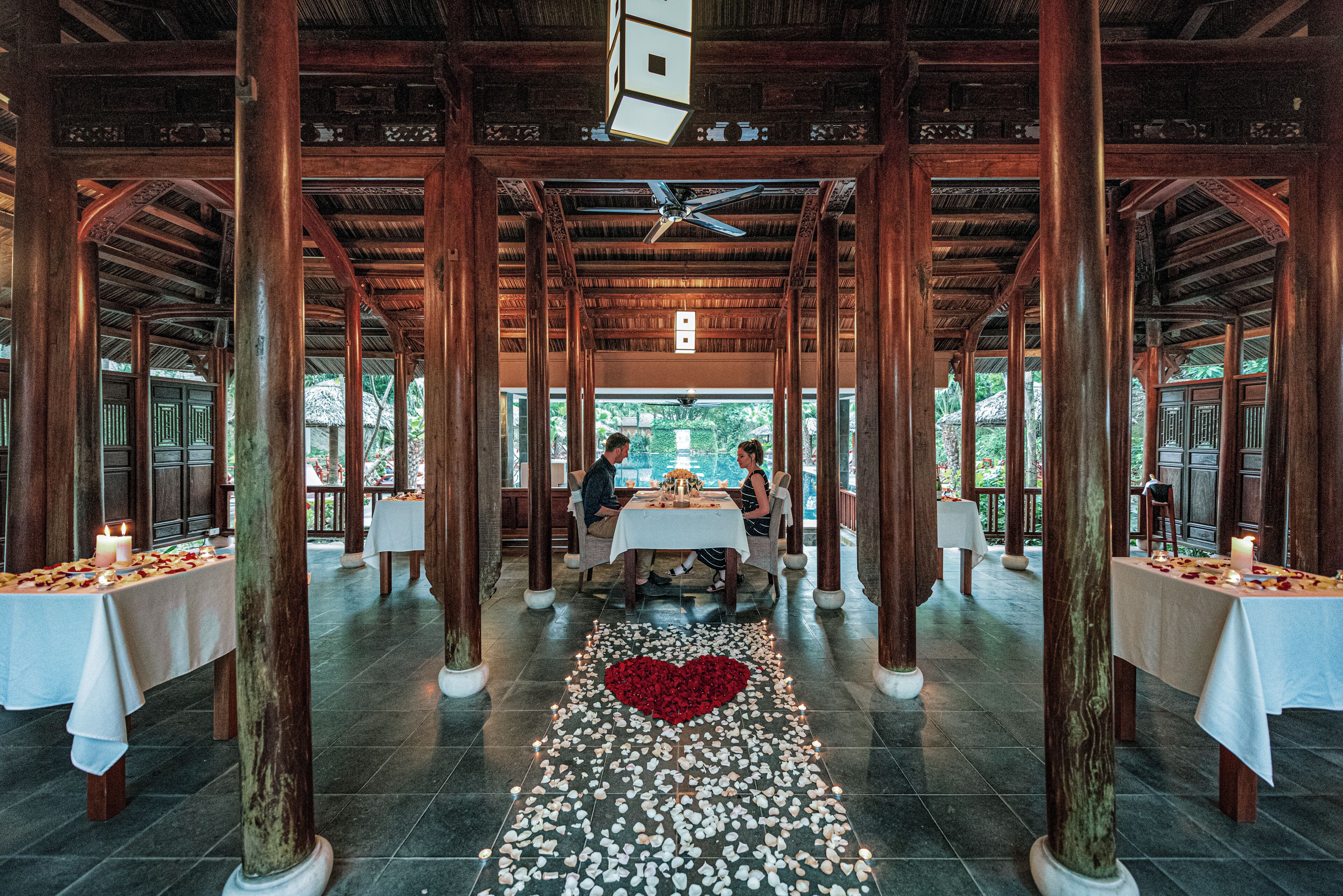 A restaurant with wooden pillars, tables with white tablecloths, and a heart-shaped arrangement of rose petals on the floor.