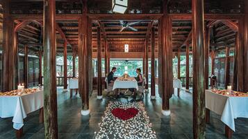 A restaurant with wooden pillars, tables with white tablecloths, and a heart-shaped arrangement of rose petals on the floor.