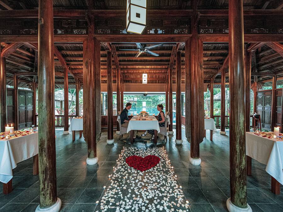 A restaurant with wooden pillars, tables with white tablecloths, and a heart-shaped arrangement of rose petals on the floor.