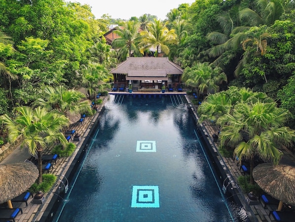 Una piscina rectangular con dos marcas azules cuadradas, rodeada de exuberante vegetación y palmeras, con un pequeño edificio al fondo.