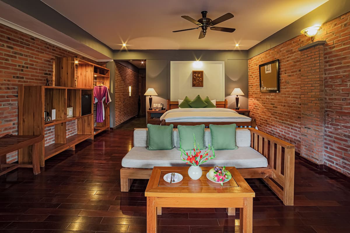 A hotel room with a brick wall, a wooden bench, a sofa, a bed with green pillows, a ceiling fan, and a nightstand with a lamp.