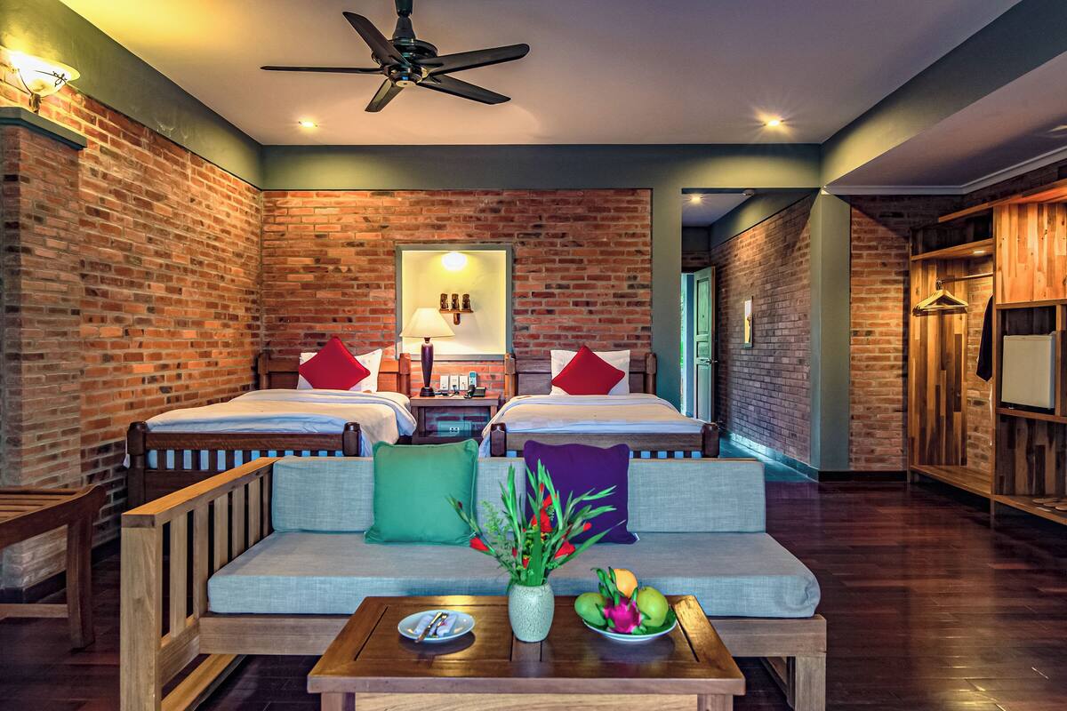A hotel room with two beds, a sofa, a table with a vase and fruit, and brick walls.