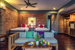 A hotel room with two beds, a sofa, a table with a vase and fruit, and brick walls.
