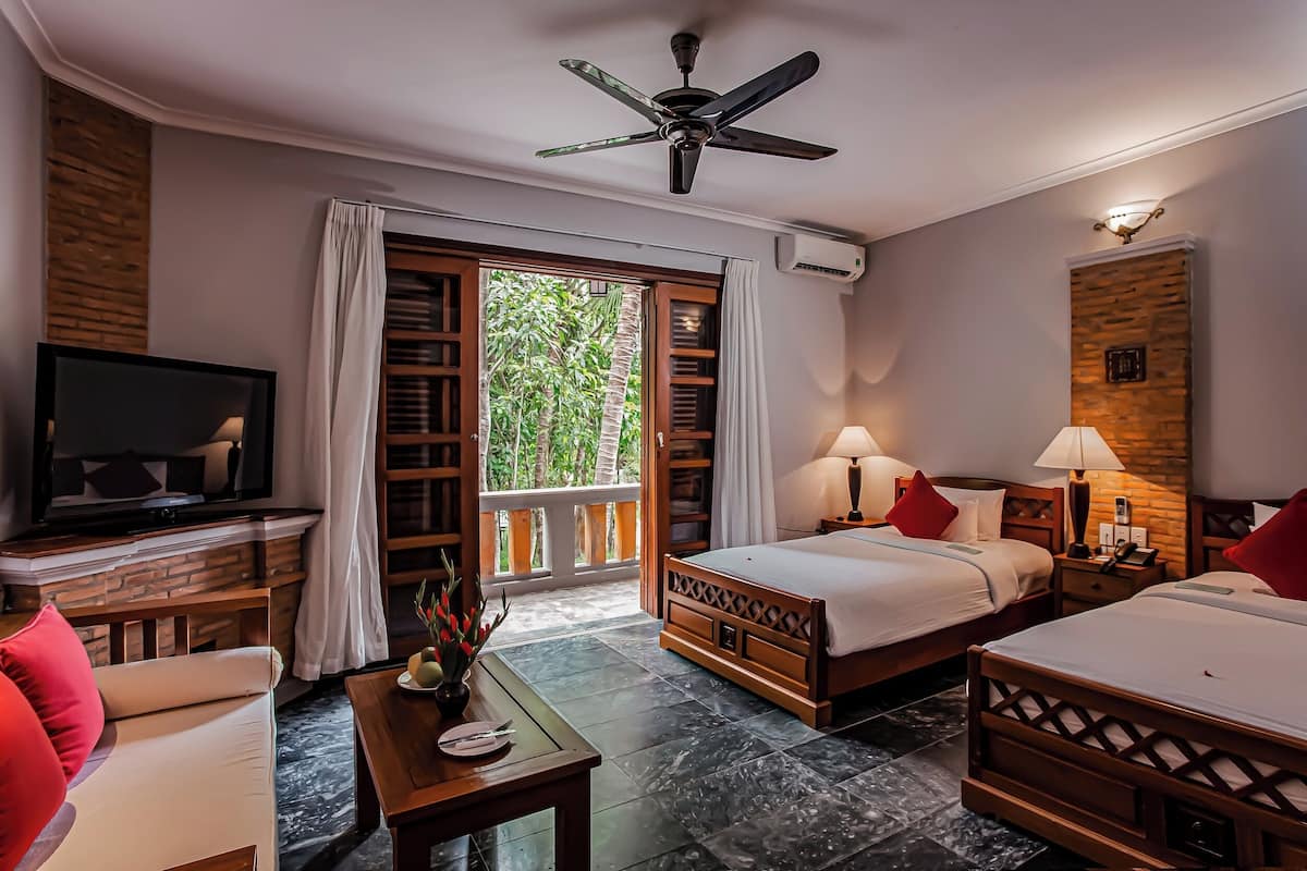 A hotel room with a TV, two beds, a sofa, a table with a vase, and a balcony with a view of greenery.