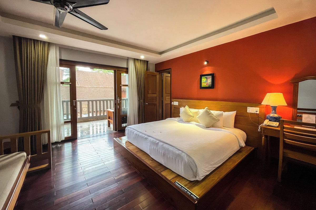 A hotel room with a bed, a desk with a lamp, a ceiling fan, and a view through a door to an outdoor area.