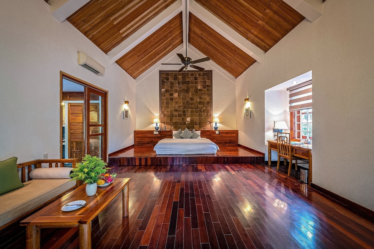 A bedroom with a sloped ceiling, a bed, a wooden desk, and a dining area.