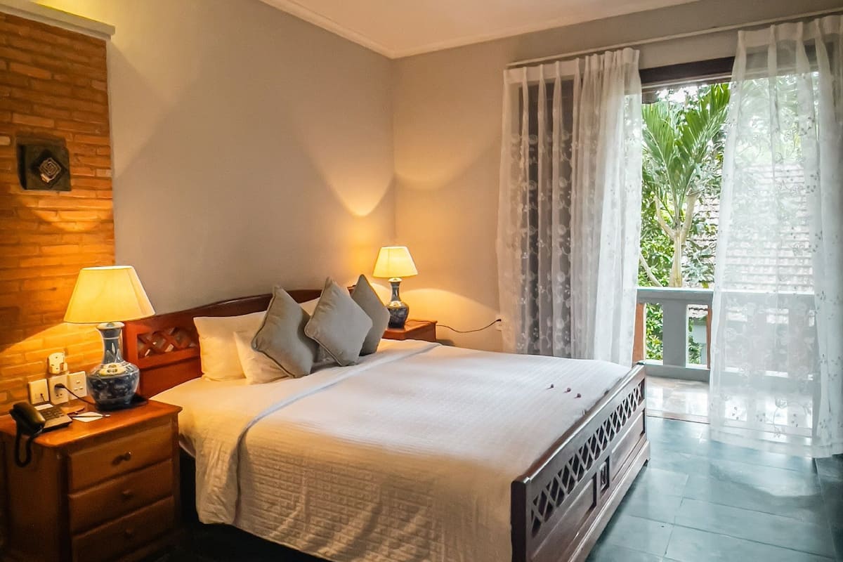 A bedroom with a bed, two bedside tables with lamps, a wooden headboard, a nightstand, and a view of greenery through the window.