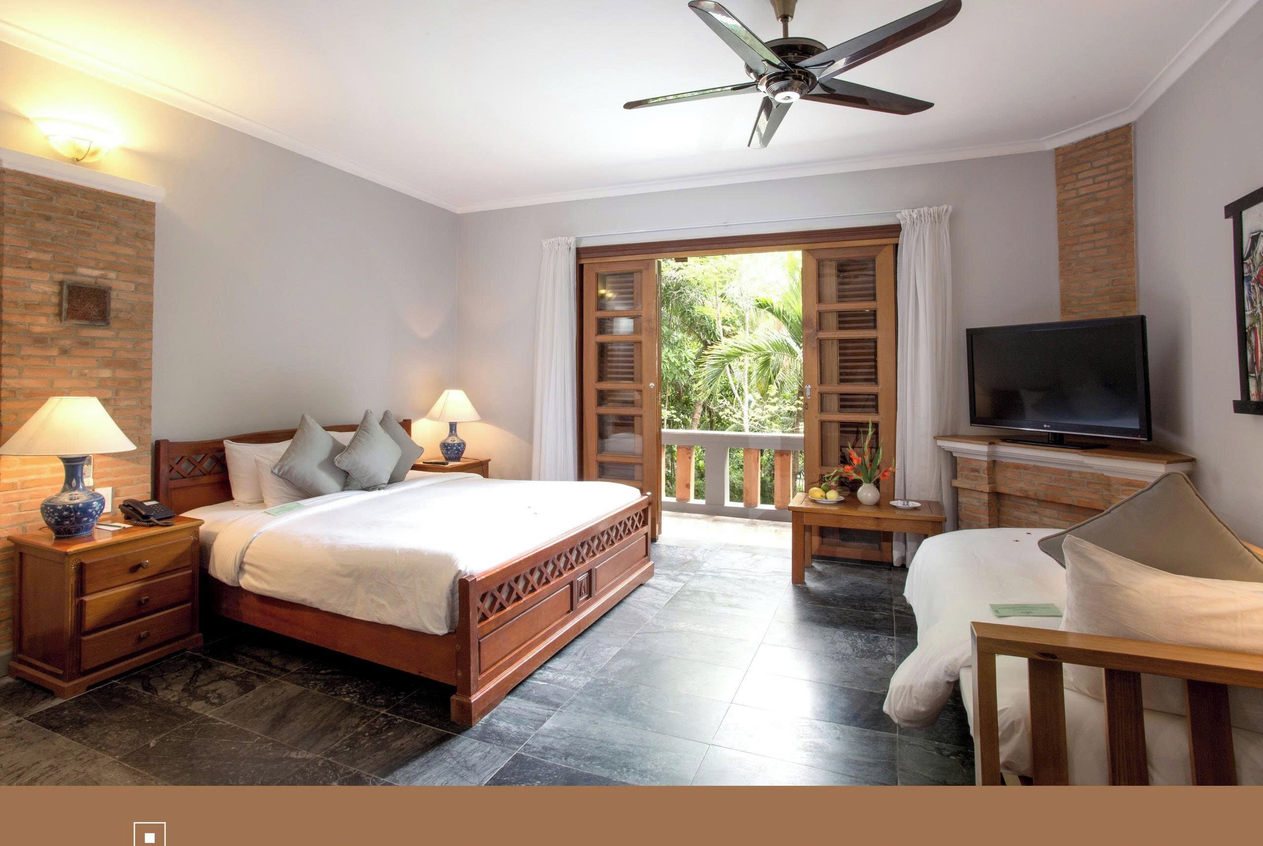 Una habitación de hotel con una cama, dos mesitas de noche con lámparas, un televisor, un balcón con vistas a la vegetación y un ventilador de techo.
