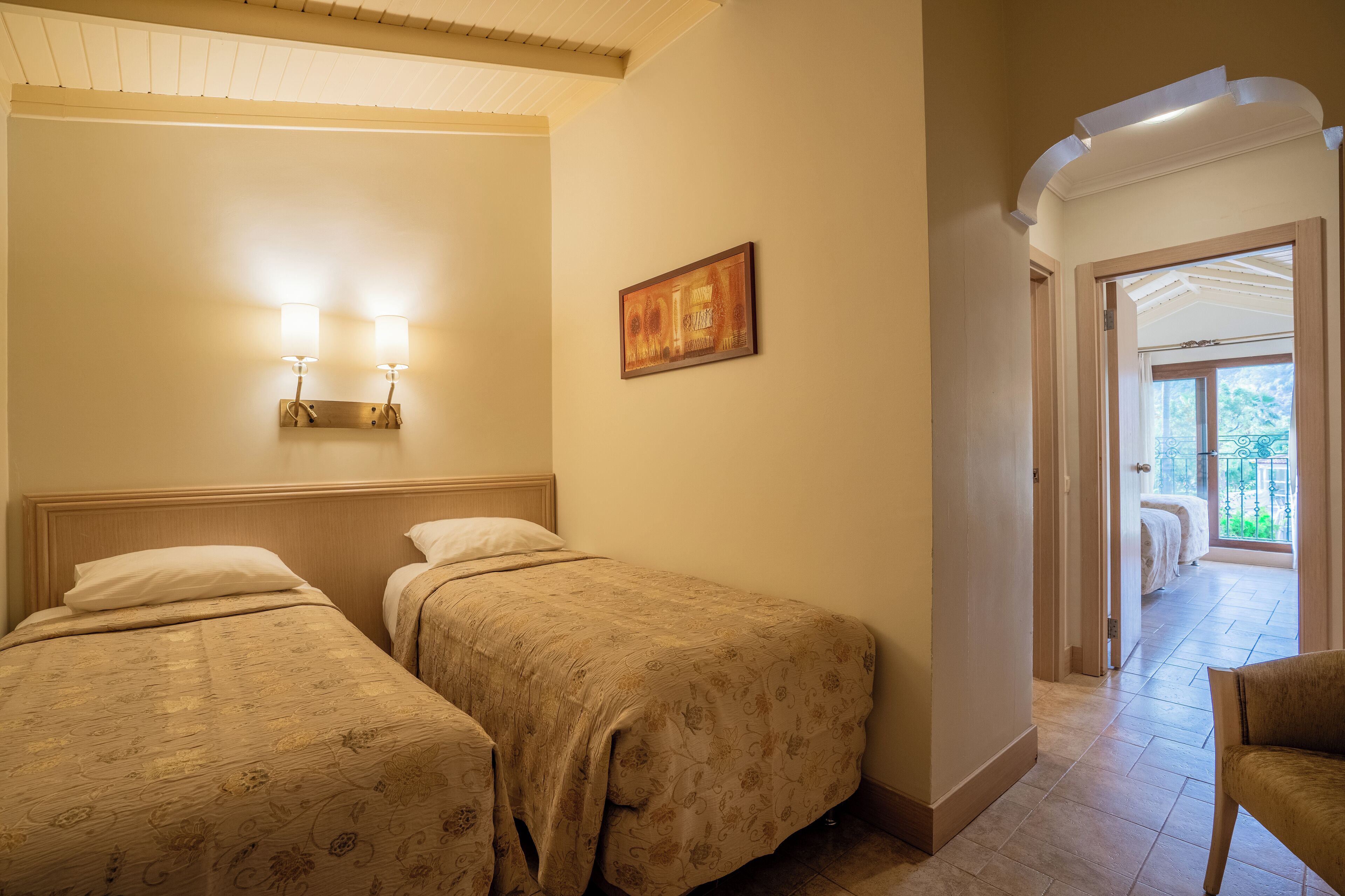 A hotel room with two beds, a painting on the wall, and a view of the outdoor balcony through an open door.