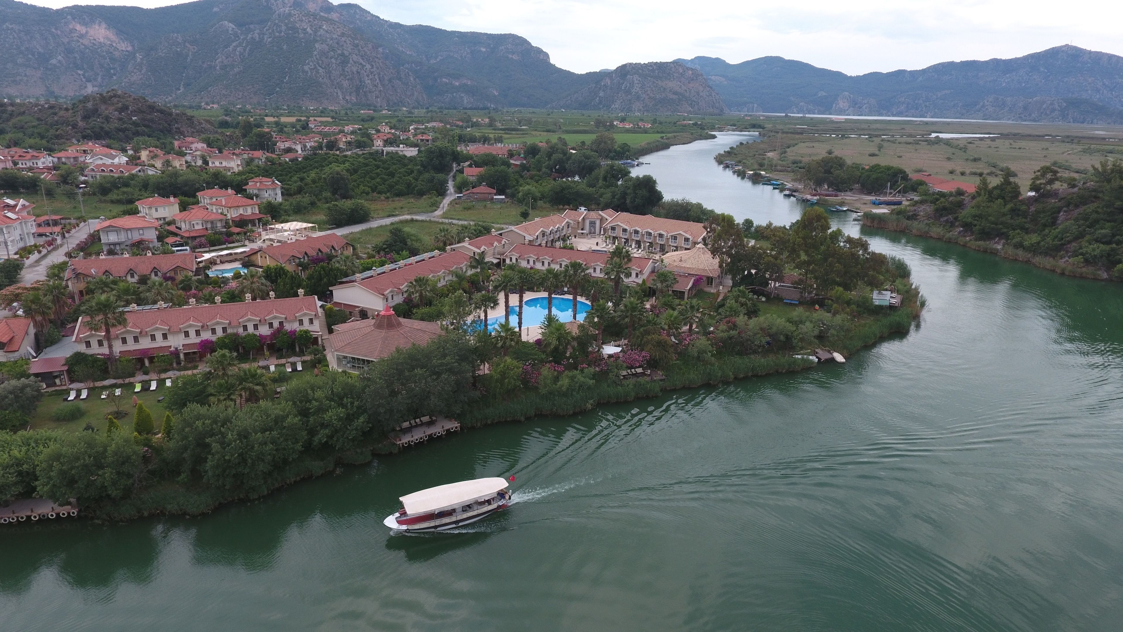 A resort with a swimming pool surrounded by buildings, a boat on the water, and mountains in the background.