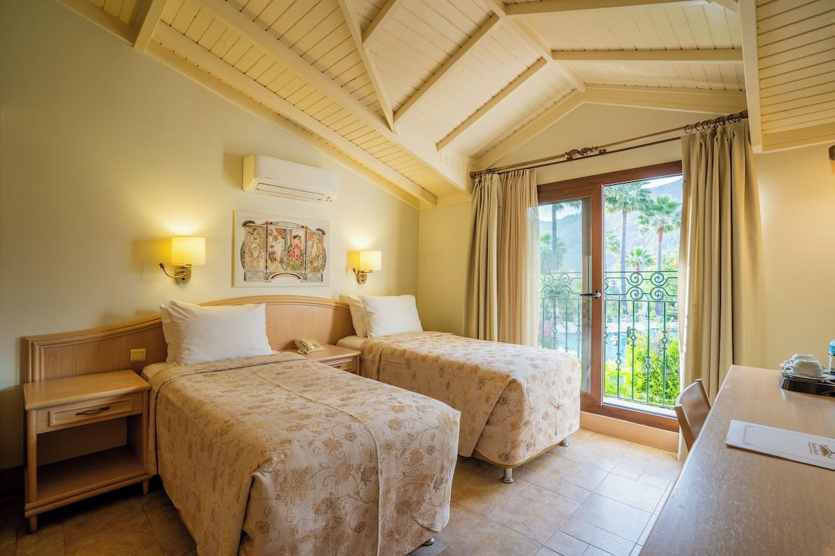 A hotel room with two beds, a wooden ceiling, a balcony with a view of palm trees, and a dining table.