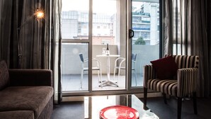 A modern living room with a sofa, a striped armchair, and a glass door leading to a balcony with a table and chairs. - Adara Hotel Richmond (Richmond)