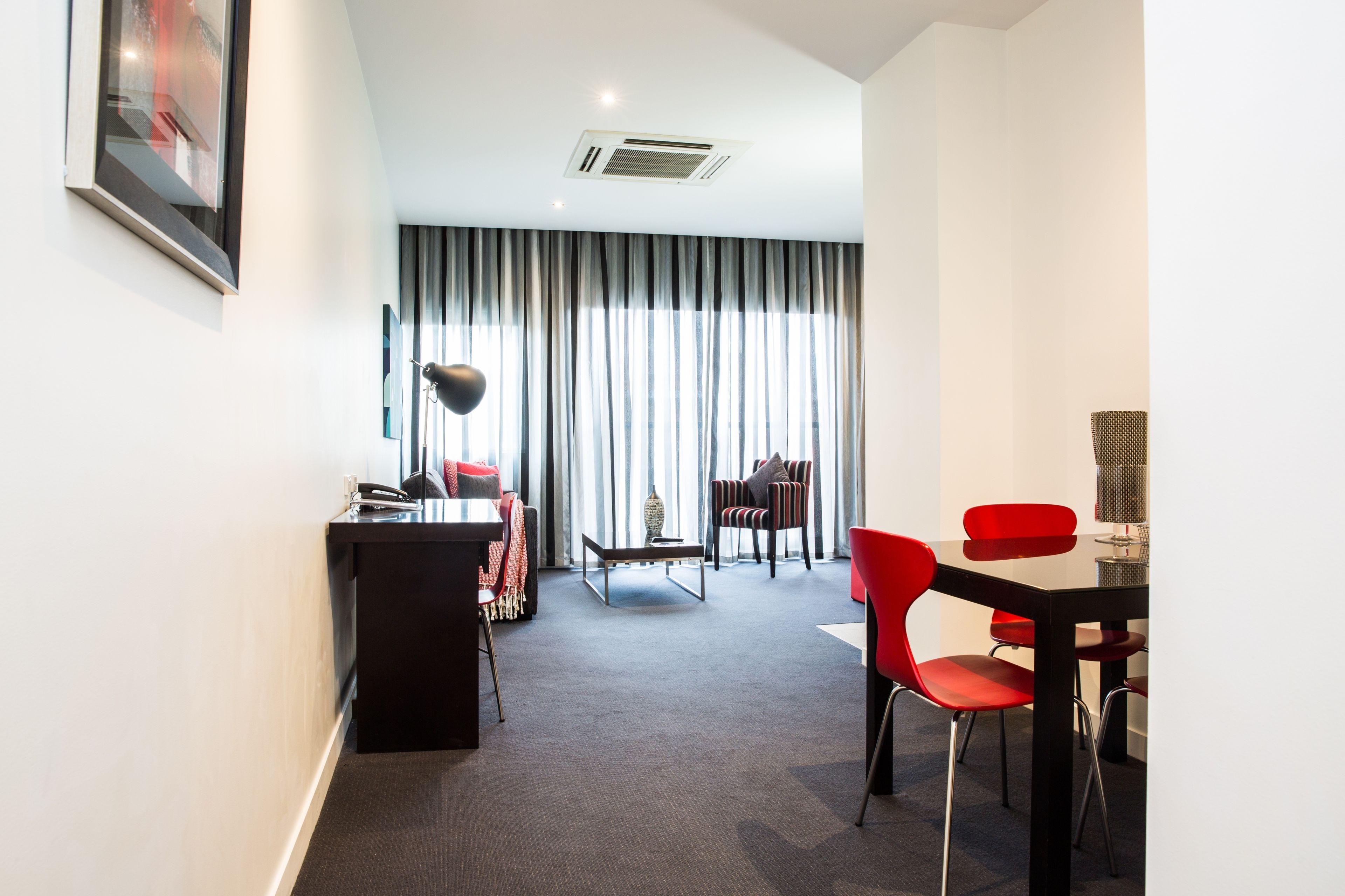 A modern hotel room with a desk, red chairs, a dining table, and a large window with curtains.