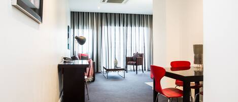 A modern hotel room with a desk, red chairs, a dining table, and a large window with curtains.